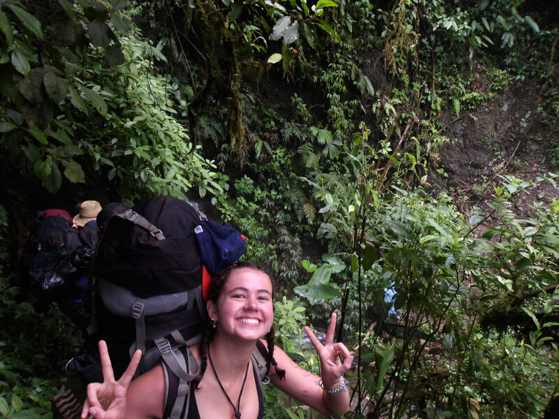 A young woman with a large backpack smiles and flashes a peace sign while hiking in a lush, green forest. She is surrounded by dense vegetation, including various plants and trees. The trail appears to be somewhat steep and rugged. She seems happy and energetic despite the challenging terrain.